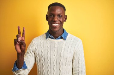 İzole sarı arka plan üzerinde kot gömlek ve beyaz kazak giyen Afrikalı Amerikalı adam gösteren ve parmaklar iki sayı ile işaret kadar emin ve mutlu gülümseyen.