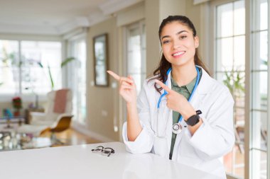 Genç doktor kadın tıbbi ceket giyen klinikte gülümseyen ve kamera iki el ve parmak tarafına işaret bakıyor.