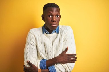 Yüzünde üzücü ve şok ifade ile kış soğuk için sallayarak ve donma izole sarı arka plan üzerinde denim gömlek ve beyaz kazak giyen Afrikalı Amerikalı adam