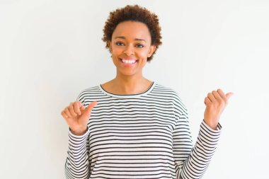 Beyaz arka planda çizgili kazak giyen genç güzel Afrikalı Amerikalı kadın el ve başparmak kadar arka arkasında Işaret, gülümseyen emin