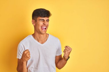 Beyaz tişört giyen genç hintli adam izole sarı arka plan üzerinde duran çok mutlu ve heyecanlı kolları kaldırdı ile kazanan jest yapıyor, gülümseyerek ve başarı için çığlık. Kutlama konsepti.