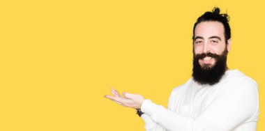 Uzun saç ve sakal sportif sweatshirt giyen genç hipster adamla el ve reklam mutlu ve kendinden emin gülümseyen sunulması avuç, yanında işaret