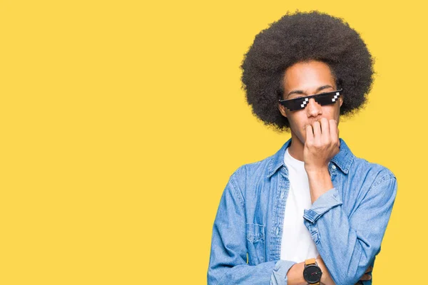 Young african american man with afro hair wearing thug life glasses looking stressed and nervous with hands on mouth biting nails. Anxiety problem.