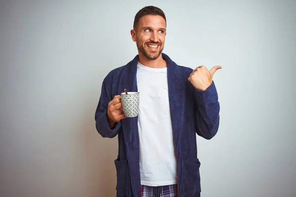 Man wearing comfortable pajamas and robe drinking cup of coffee over isolated background with angry face, negative sign showing dislike with thumbs down, rejection concept