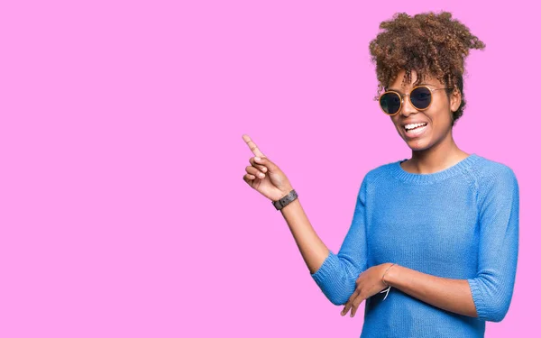 Beautiful young african american woman wearing sunglasses over isolated background with a big smile on face, pointing with hand and finger to the side looking at the camera.