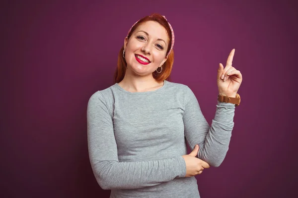 美丽的红发女人穿着灰色T恤 在孤立的紫色背景上 脸上带着灿烂的笑容 用手和手指指着一边看着相机 — 图库照片