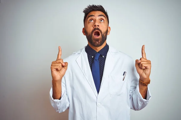 Young indian doctor man standing over isolated white background amazed and surprised looking up and pointing with fingers and raised arms.