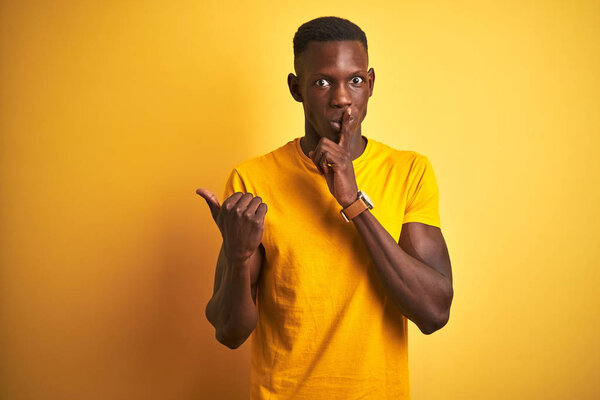 Young african american man wearing casual t-shirt standing over isolated yellow background asking to be quiet with finger on lips pointing with hand to the side. Silence and secret concept.