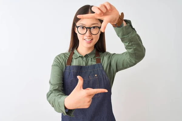 エプロンとメガネをかけた中国の若い店主女性が 幸せそうな顔をした手と指でフレームを作る孤立した白い背景の上に笑顔 創造性と写真のコンセプト — ストック写真