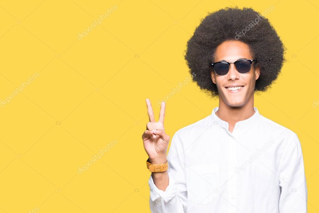 Young african american man with afro hair wearing sunglasses smiling with happy face winking at the camera doing victory sign. Number two.