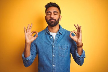 İzole sarı arka plan üzerinde ayakta kot gömlek giyen genç Hint adam rahatlayın ve gözleri ile gülümseyen meditasyon jest parmaklar ile kapalı. Yoga konsepti.