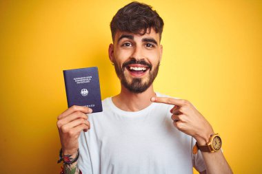 Dövme ile genç adam izole sarı arka plan üzerinde Alman Almanya pasaportu giyiyor el ve parmak ile işaret çok mutlu