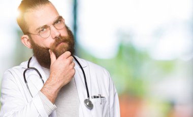 Sakal ile genç sarışın doktor adam el çene hakkında soru, dalgın ifade düşünme ile tıbbi ceket giyiyor. Düşünceli bir yüz ile gülümsüyor. Şüphe kavramı.