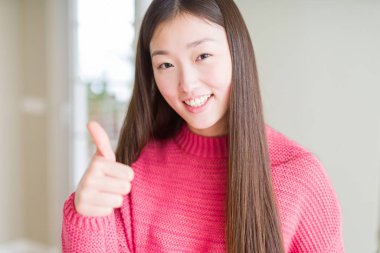 Pembe kazak giyen güzel Asyalı kadın el ile jest kadar mutlu başparmak yapıyor. Başarı gösteren kameraya bakarak ifadeyi onaylama.