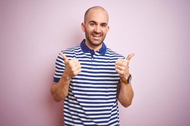 Sakal giyen rahat çizgili mavi t-shirt üzerinde pembe izole arka plan başarı işareti ile genç kel adam el ile olumlu jest yapıyor, gülümseyen ve mutlu yukarı başparmak. Neşeli ifade ve kazanan jest.