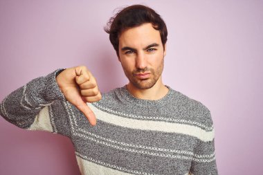 Kızgın yüz ile izole pembe arka plan üzerinde ayakta Casual çizgili kazak duran genç yakışıklı adam, aşağı başparmak ile nefret gösteren negatif işareti, ret kavramı
