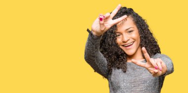 Kıvırcık saçlı giyen ile genç güzel kadın gri kazak zafer işareti yapıyor parmak gösterilen kamera seyir gülümseyen. İki numaralı.