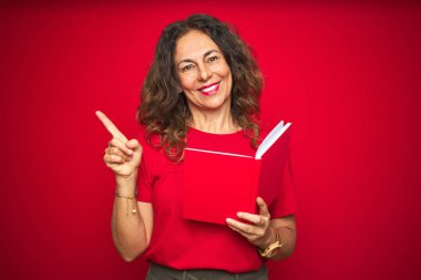Orta yaş üst düzey kadın yan el ve parmak ile işaret çok mutlu kırmızı izole arka plan üzerinde bir kitap okuma