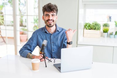 Genç adam kayıt podcast mikrofon ve dizüstü mutlu yüz görünümlü ile gülümseyen ve başparmak ile yan işaret kullanarak.