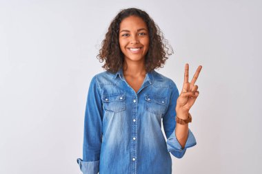 Genç Brezilyalı kadın, izole beyaz arka plan üzerinde ayakta kot gömlek giymiş ve parmaklar iki numara ile işaret kadar emin ve mutlu gülümseyen.