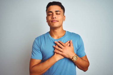 Genç Brezilyalı adam kapalı gözler ve yüz minnettar jest ile göğsüne eller ile gülümseyen izole beyaz arka plan üzerinde ayakta mavi t-shirt giyiyor. Sağlık konsepti.