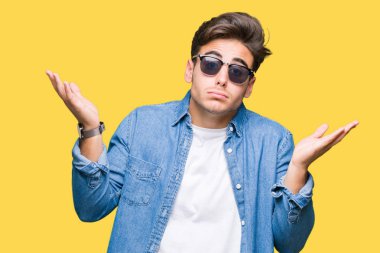 Genç yakışıklı adam izole arka plan clueless ve şaşkın bir ifadeyle kollar ve eller kaldırdı üzerinde güneş gözlüğü takıyor. Şüphe kavramı.