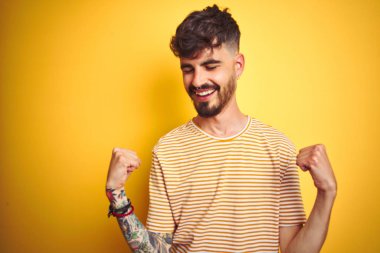 Dövme ile genç adam izole sarı arka plan üzerinde duran çizgili t-shirt ayakta sürpriz kutluyor ve silah ile başarı için hayran kaldırdı ve gözler kapalı. Kazanan kavramı.