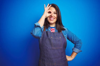 Mavi izole arka plan üzerinde mağaza üniforma apron giyen genç güzel iş kadını el gülümseyen ile ok jest yapıyor, göz parmaklarıyla mutlu yüz görünümlü.