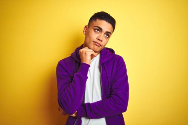 Genç Brezilyalı adam, çene üzerinde el ile izole sarı arka plan üzerinde ayakta mor sweatshirt giyen soru hakkında düşünme, dalgın ifade. Düşünceli bir yüz ile gülümsüyor. Şüphe kavramı.