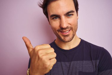 Genç yakışıklı adam izole pembe arka plan üzerinde ayakta rahat t-shirt giymiş büyük bir gülümseme ile ok işareti, parmak yukarı başparmak, mükemmel işareti yapıyor mutlu