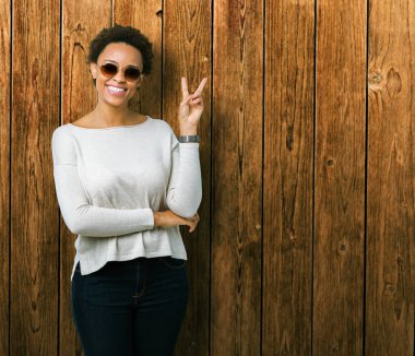 Güzel genç Afro-Amerikan kadın mutlu yüz zafer işareti yaparken kameraya göz kırpıyor ile gülümseyen izole arka plan üzerinde güneş gözlüğü takıyor. İki numaralı.