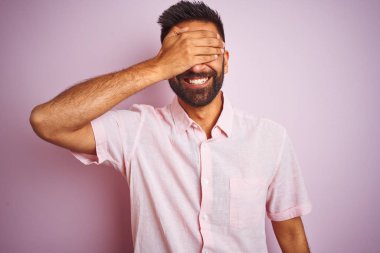 Genç Hintli adam rahat gömlek ayakta izole pembe arka plan gülümseyen ve sürpriz için gözleri kapsayan yüzünde el ile gülüyor giymiş. Kör kavram.