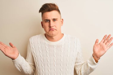 Genç yakışıklı adam kış kazak giyen izole beyaz arka plan üzerinde duran habersiz ve kolları ve elleri kaldırdı ile karışık ifade. Şüphe kavramı.