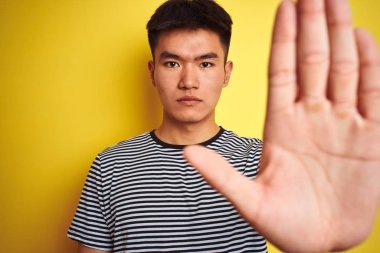 Açık el ile izole sarı arka plan üzerinde duran çizgili t-shirt ayakta giyen genç Asyalı Çinli adam ciddi ve emin ifade ile stop işareti yapıyor, savunma jest