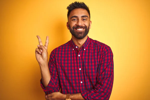 Young indian man wearing casual shirt standing over isolated yellow background smiling with happy face winking at the camera doing victory sign. Number two.