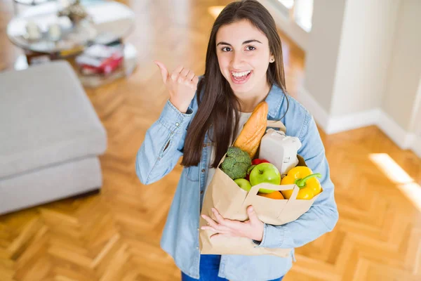 健康的な食料品でいっぱいの紙袋を持つ美しい若い女性は 幸せそうな顔で横に親指で示しています — ストック写真