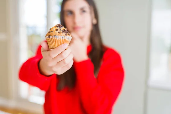 Beautiful young woman eating chocolate chips muffin serious face thinking about question, very confused idea