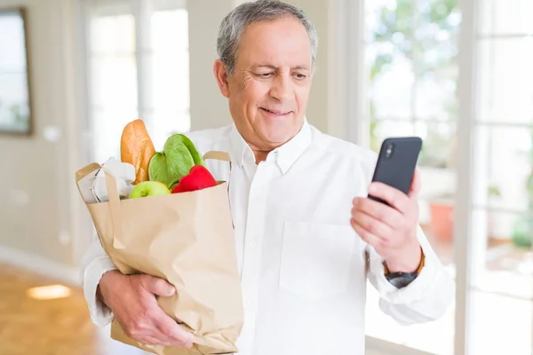 Handsome senior man holding paper bag full of fresh groceries and looking at smartphone smiling
