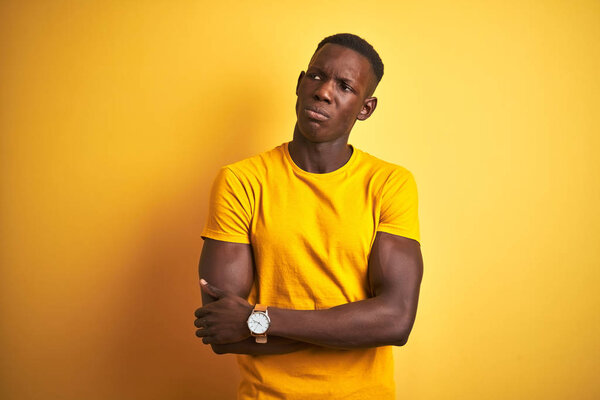 Young african american man wearing casual t-shirt standing over isolated yellow background looking to the side with arms crossed convinced and confident