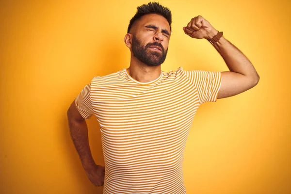 Young indian man wearing t-shirt standing over isolated yellow background stretching back, tired and relaxed, sleepy and yawning for early morning