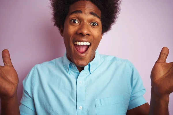 Young american man with afro hair wearing blue shirt standing over isolated pink background very happy and excited, winner expression celebrating victory screaming with big smile and raised hands