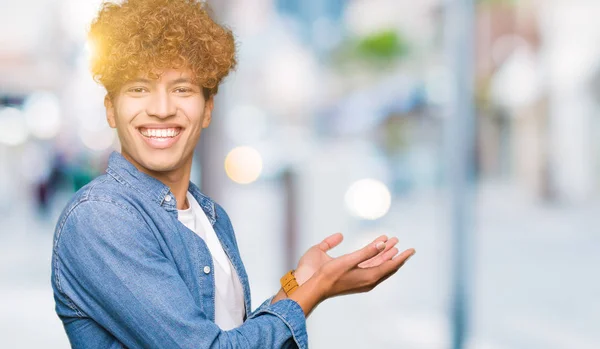 Young handsome man with afro hair wearing denim jacket Pointing to the side with hand and open palm, presenting ad smiling happy and confident