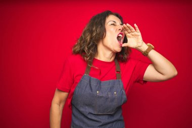 Orta yaş üst düzey kadın kırmızı izole arka plan üzerinde apron üniforma giyiyor ve ağzına el ile yan gürültülü çığlık. İletişim konsepti.