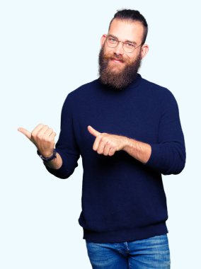 Gözlük ve balıkçı yaka kazak giyen genç sarışın adam kendinden emin gülümseyen arkasından el ve kadar başparmak ile işaret