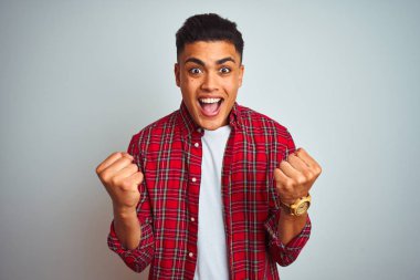 İzole beyaz arka plan üzerinde ayakta kırmızı gömlek giyen genç Brezilyalı adam şaşırttı ve silah kaldırdı ve açık gözleri ile başarı için hayran kutluyor. Kazanan kavramı.