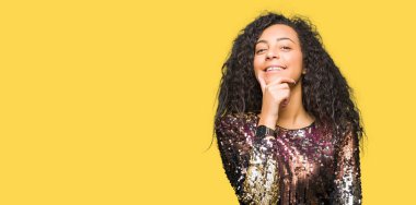 Kıvırcık saçlı emin çapraz kollar ve çeneme kaldırdı el ile gülümseme ile kameraya bakarak gece parti elbise genç güzel kız. Pozitif düşünce.