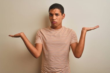 Genç yakışıklı Arap adam izole beyaz arka plan üzerinde çizgili t-shirt giyen habersiz ve kollar ve eller kaldırdı ile karışık ifade. Şüphe kavramı.