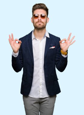 Genç yakışıklı zarif adam giyen güneş gözlüğü ve moda ceket dinlenmek ve gülümseyen gözlerle parmak ile meditasyon jest yapıyor kapalı. Yoga kavramı.