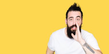 Uzun saç ve sakal ile genç hipster adam rahat beyaz t-shirt el ağzına gizli söylenti söyleyerek, kötü niyetli konuşma konuşma fısıldayan giyiyor