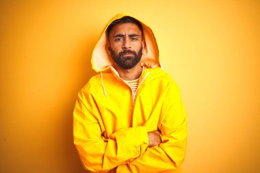 İzole sarı arka plan üzerinde Hood ayakta ile yağmurluk giyen genç Hintli adam şüpheci ve sinir, Çapraz kollar ile yüz üzerinde ifade onaylamıyor. Negatif kişi.